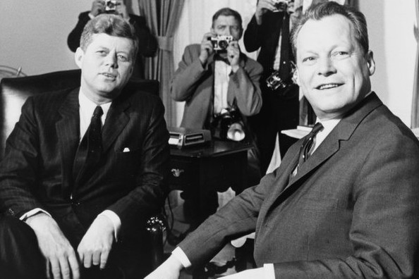 Schwarz-Weiß-Aufnahme von Willy Brandt und John F. Kennedy. Hinter den beiden sind 3 Fotografen zu sehen, die den Finger bereits am Auslöser haben.