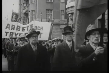 Schwarz-Weiß-Aufnahme aus der Schwedischen Wochenschau von der Ersten Mai-Kundgebung im Jahr 1944. Im Hintergrund sieht man eine Marschkapelle und ein Transparent mit der Aufschrift: „Arbete och trygghet“.,