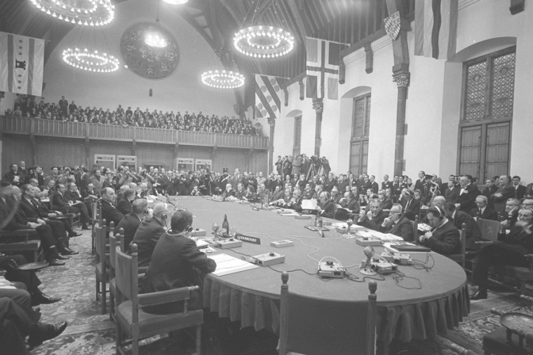Schwarz-Weiß-Aufnahme aus dem Jahr 1969 von einem Gipfeltreffen der Europäischen Wirtschaftsgemeinschaft. Dabei sitzen die Staats- und Regierungschefs an einem ovalen Tisch zusammen. Hinter Ihnen eine Vielzahl von Beamten. Auch auf der Tribüne im hinteren Teil des Saales stehen viele Menschen.