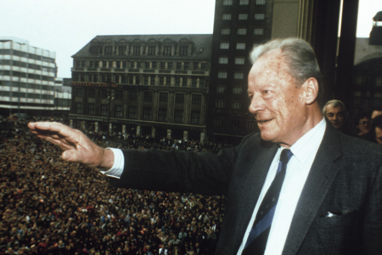 Willy Brandt steht auf einem Balkon und winkt der Menschenmasse vor Ihm auf dem Platz zu.