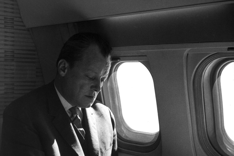 Schwarz-Weiß-Aufnahme von Willy Brandt, der in einem Flugzeug sitzt und mit dem Blick lesend nach unten schaut.