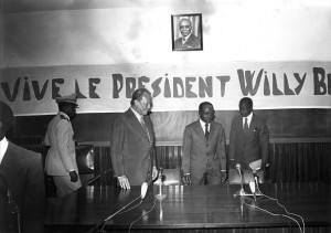 Schwarz-Weiß-Aufnahme von Willy Brandt bei einer Pressekonferenz mit dem senegalesischen Präsidenten. Im Hintergrund ist ein Transparent zu sehen, auf dem steht: „Vive le president Willy Brandt“.
