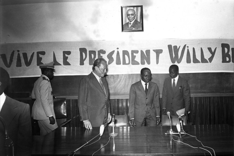 Schwarz-Weiß-Aufnahme von Willy Brandt bei einer Pressekonferenz mit dem senegalesischen Präsidenten. Im Hintergrund ist ein Transparent zu sehen, auf dem steht: „Vive le president Willy Brandt“.