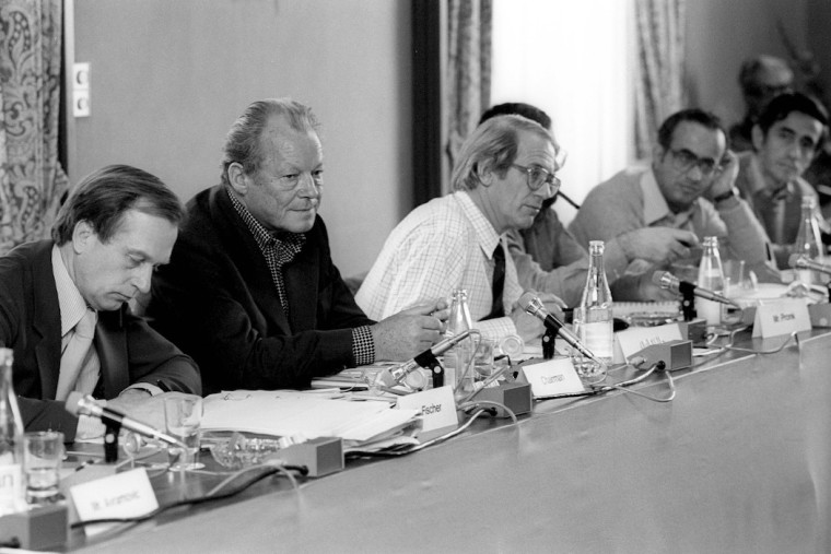 Willy Brandt sitzt neben Fritz Fischer, Göran Ohlin, Jan Pronk und Abdlatif Y. Al-Hamad an einem Tisch bei der Sitzung der Nord-Süd-Kommission. Fotografie in Schwarz-Weiß.