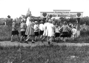 Schwarz-Weiß-Aufnahme aus dem Jahr 1927 bei der eine Gruppe von Kindern mit Spaten ausgerüstet durch ein Tor läuft, welches die Aufschrift „Ausschuss für Arbeiter Wohlfahrt Lübeck“ trägt.