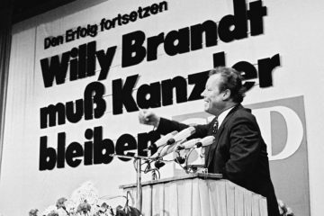 Schwarz-Weiß-Aufnahme Willy Brandts aus dem Jahre 1972 vor einem Rednerpult. Hinter Ihm eine Leinwand auf der steht: Den Erfolg fortsetzen. Willy Brandt muss Kanzler bleiben. Öffnet eine Sammlung von Zitaten Willy Brandts.