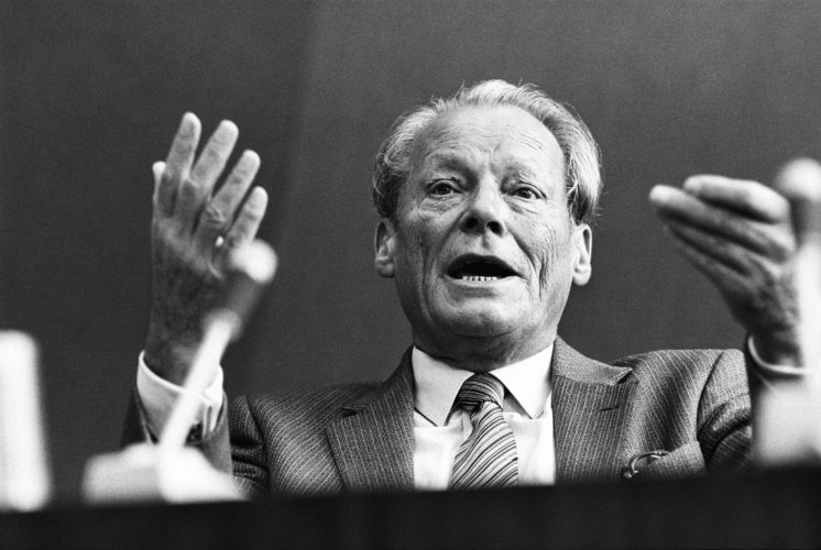 Schwarz-Weiß Porträt von Willy Brandt der gestikulierenden die Hände in die Höhe hält. Im Vordergrund ist unscharf ein Mikrofon zu sehen. Aufgenommen wurde dieses Foto 1978 bei einer Rede vor den Vereinten Nationen in New York.