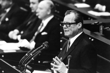 Schwarz-Weiß-Aufnahme von Willy Brandt aus dem Jahre 1969 vor einem Rednerpult im Bonner Bundestag. Links neben ihm ist unscharf auf der Regierungsbank Walter Scheel zu sehen.